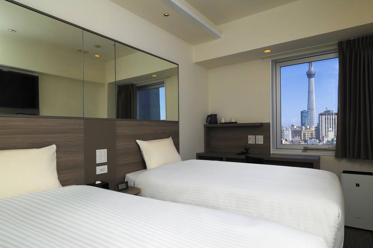 A hotel room with two beds, a large mirror, a desk with a chair, a TV, and a window with a cityscape view.