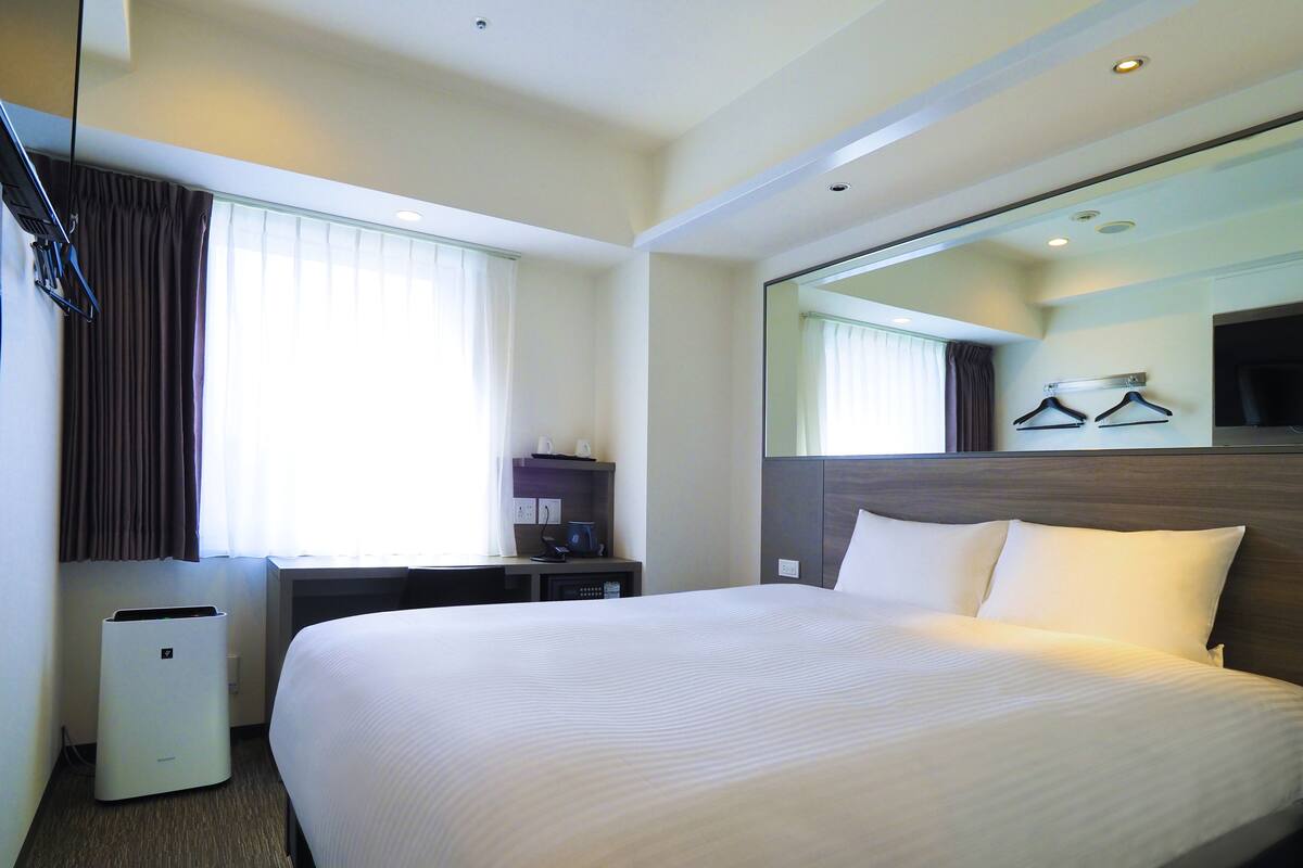 A hotel room with a large bed, a desk with a computer, a mirror, and a window with curtains.
