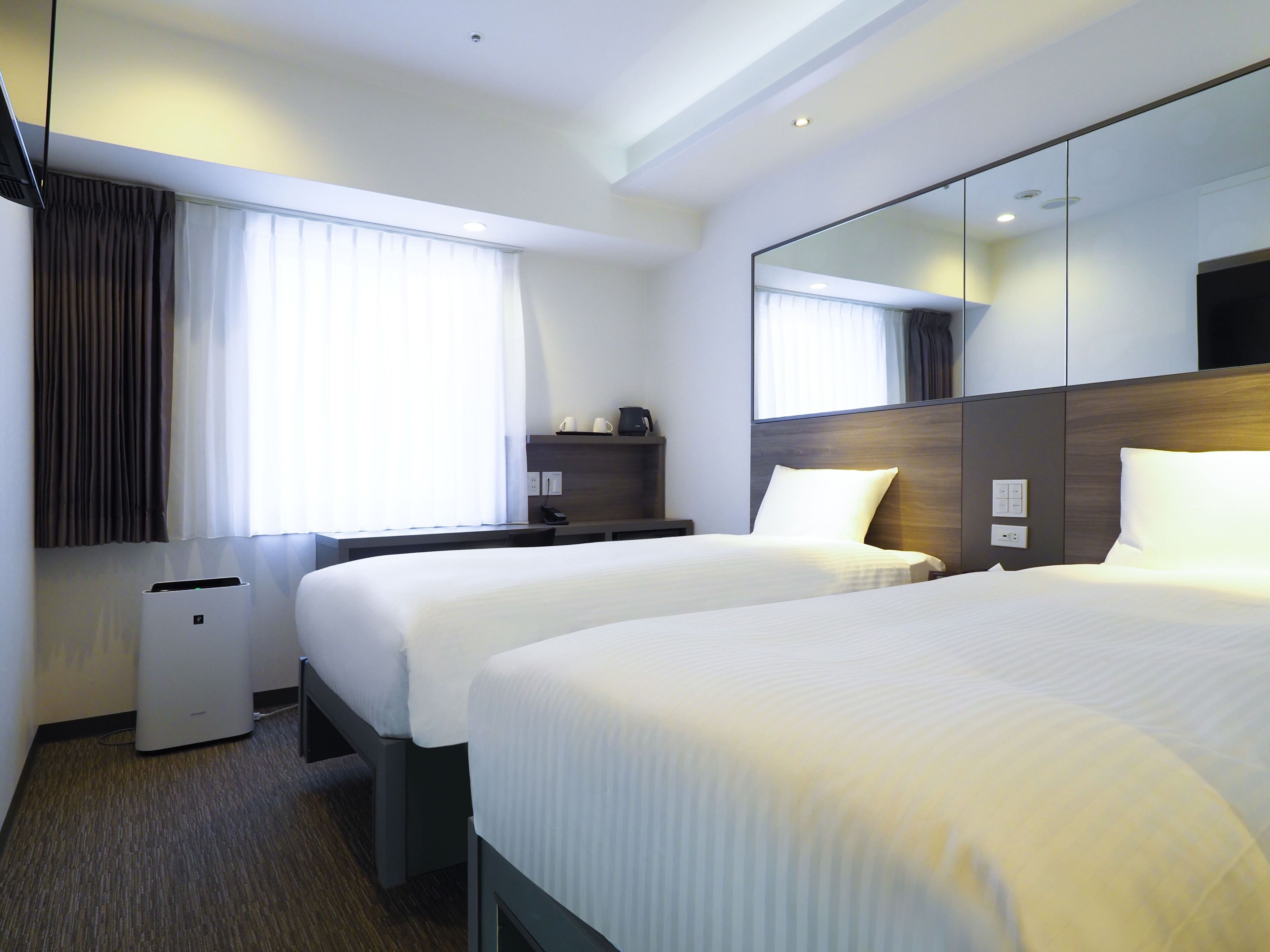 A hotel room with two beds, a desk, a mirror, and a window with curtains.