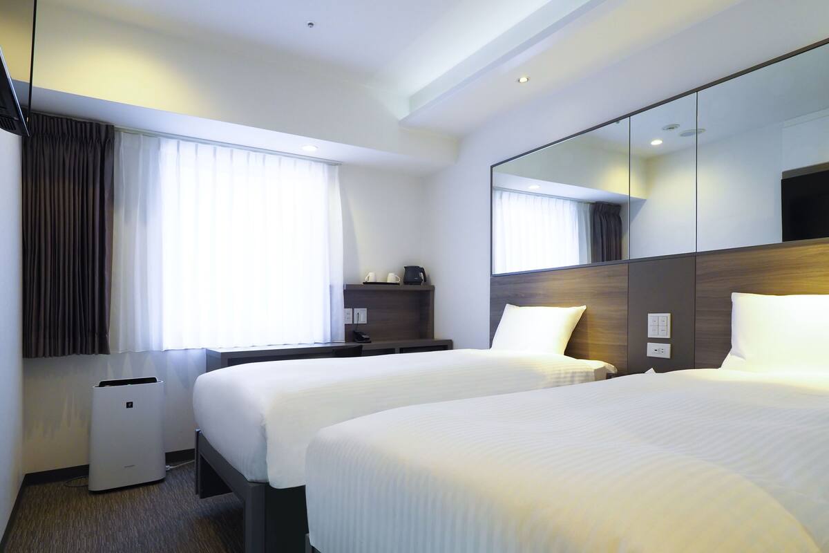 A hotel room with two beds, a desk, a mirror, and a window with curtains.