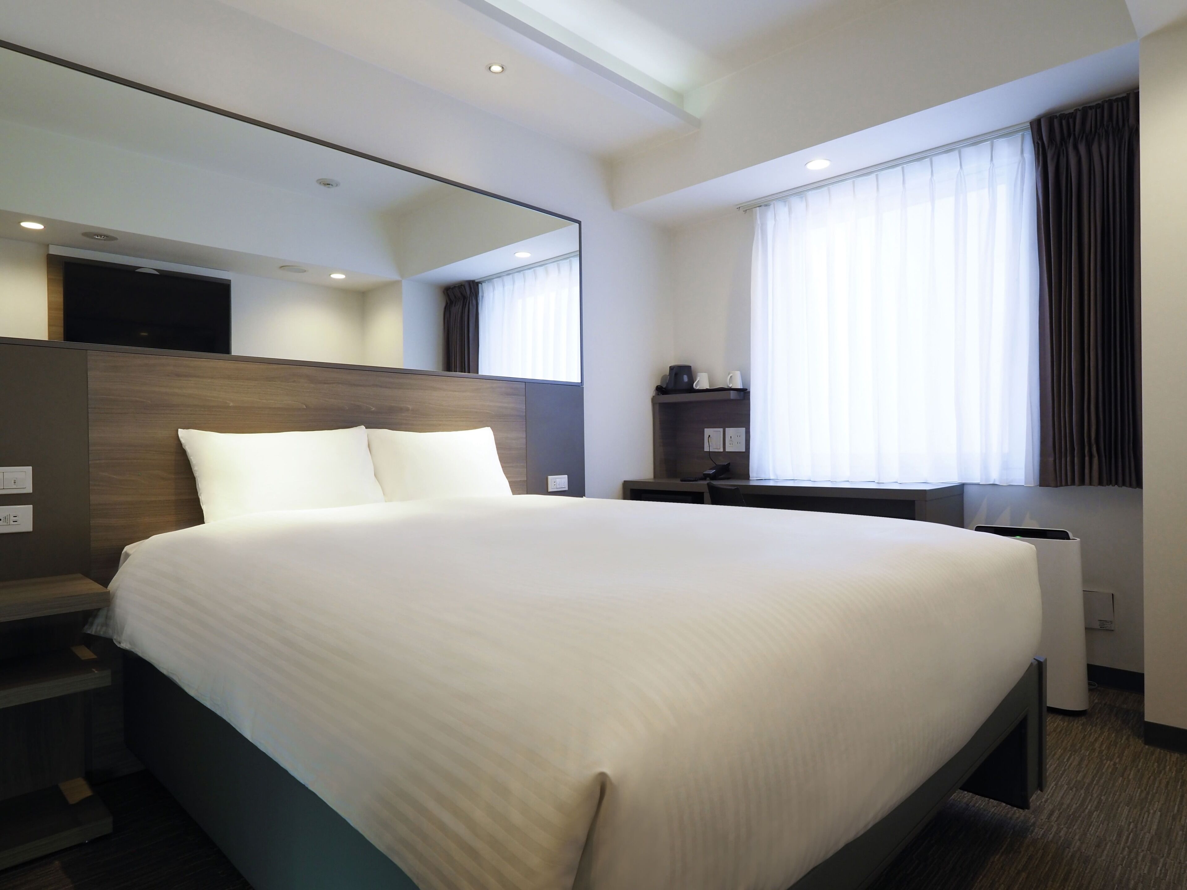 A modern hotel room with a large bed, a bedside table, a desk, a chair, a mirror, and a window with curtains.