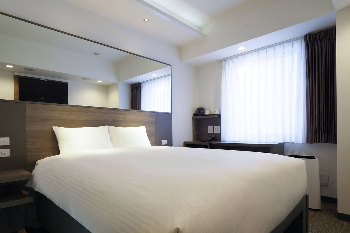 A modern hotel room with a large bed, a bedside table, a desk, a chair, a mirror, and a window with curtains.
