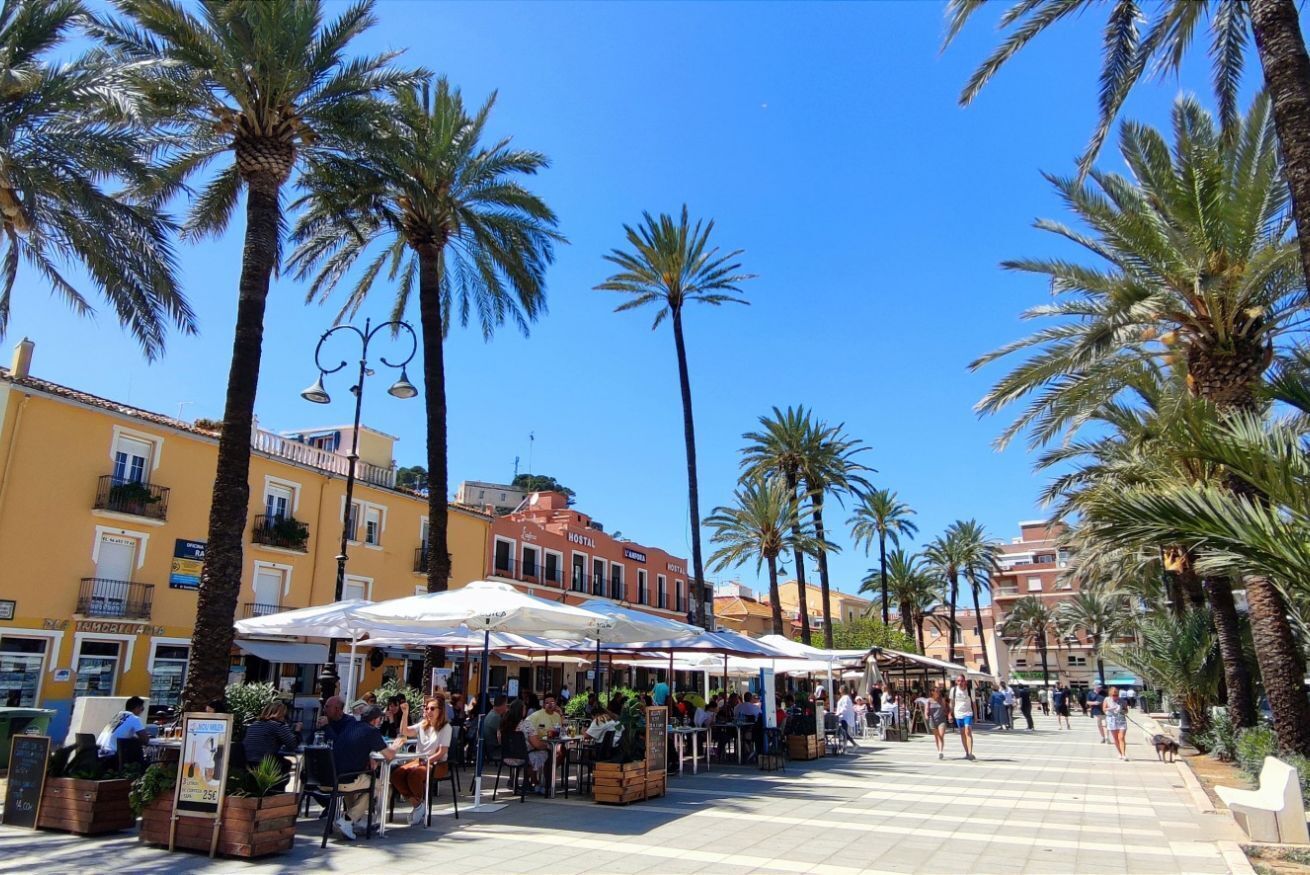 A bustling outdoor café area with tables and chairs, surrounded by palm trees and buildings with balconies.
