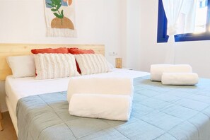 A neatly made bed with white and red pillows, a blue quilt, and two rolled towels on the floor.