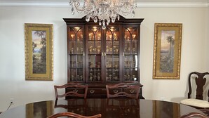 A formal dining room with a large wooden table, ornate chairs, a glass cabinet with glassware, and two framed landscape paintings. - The Bays Edge Retreat Waterfront (Bradenton)