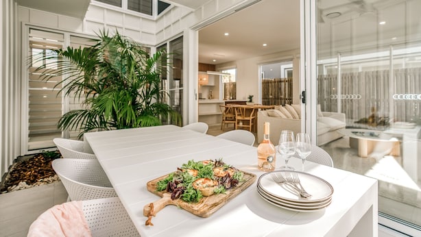 A dining area with a white table set for two, featuring a wooden serving board with a salad, a bottle of wine, and glasses.