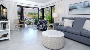 A modern living room with a grey sofa, white armchairs, a round coffee table, and a large window offering a view of palm trees. - C14 Moon Bay - Recently renovated 2 bedroom townhome (Key Largo)