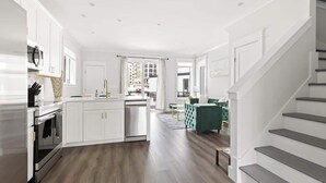 A modern kitchen with white cabinetry, a stainless steel range, and a dishwasher. There is a dining area with a green upholstered chair and a staircase leading to another level. - Inspiration at The Cove Riverwalk Villas (Wilmington)