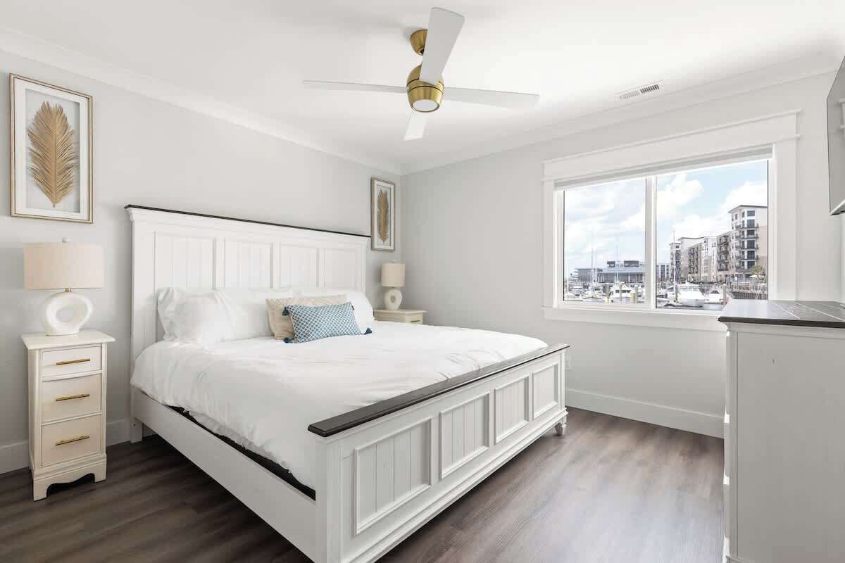 Una camera da letto con un letto bianco, una ventola a soffitto, un comodino, una lampada e una finestra con vista sulla città.