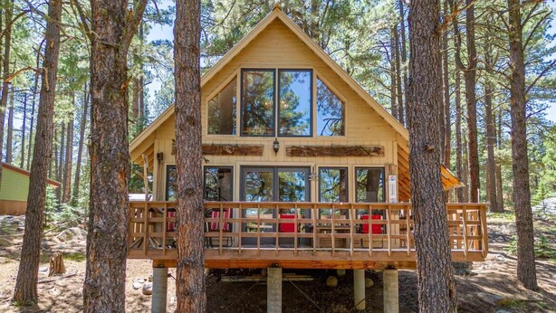 A wooden cabin with large windows, a deck with chairs, and a forest backdrop.