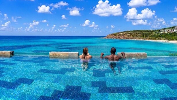 Una piscina azul y transparente con bordes de azulejos, vistas al océano y una costa lejana con edificios.
