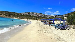 Una playa con aguas azules cristalinas, tumbonas con sombrillas azules, y una hilera de edificios al fondo.