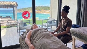Una habitación con un amplio ventanal con vistas a la playa, una camilla de masaje y un terapeuta atendiendo a un cliente.