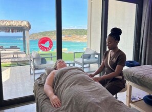 Une pièce dotée d’une grande fenêtre donnant sur une plage, d’une table de massage et d’un massothérapeute qui s’occupe d’un client.