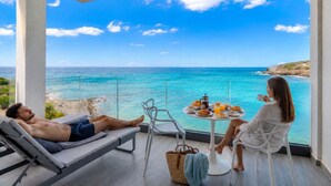 Un balcón con vista al mar, una silla de descanso, una mesa de comedor con desayuno, y una silla para dos.