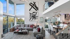 A spacious living room with a large sofa, a coffee table, and a rug. There are abstract paintings on the wall and a staircase leading to an upper level.