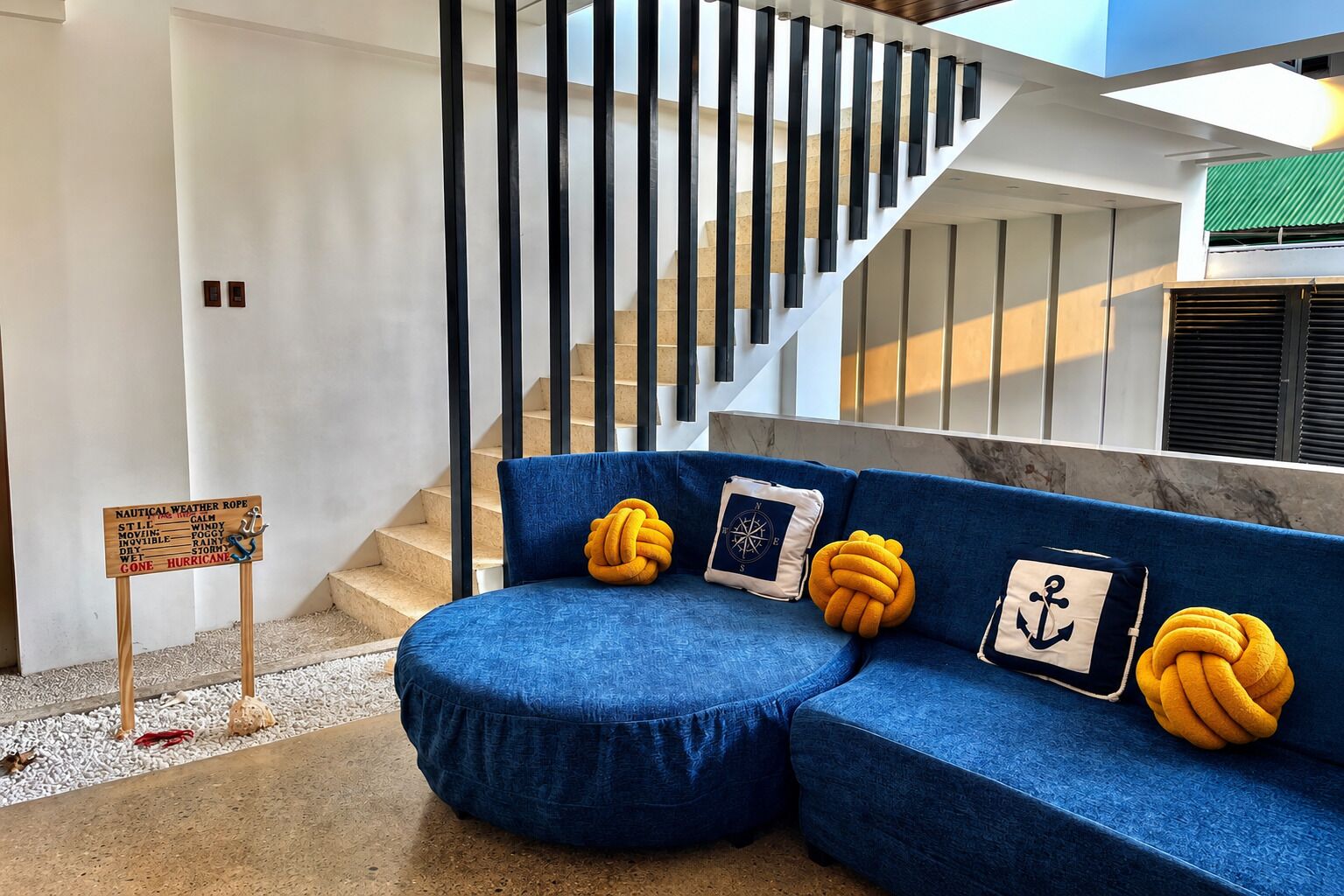A modern interior with a blue sofa, nautical-themed cushions, a weather-themed sign, and a staircase.