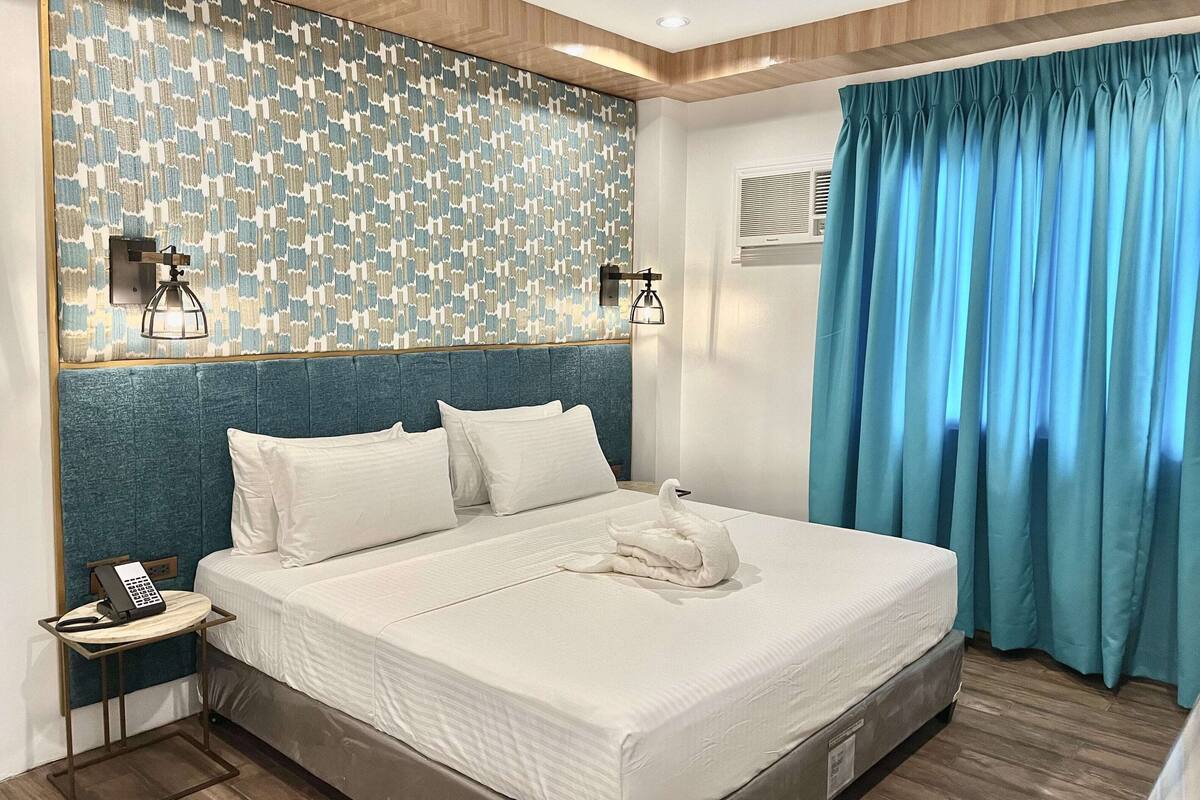 A hotel room with a bed, a telephone, a wall-mounted light fixture, and a window with curtains.