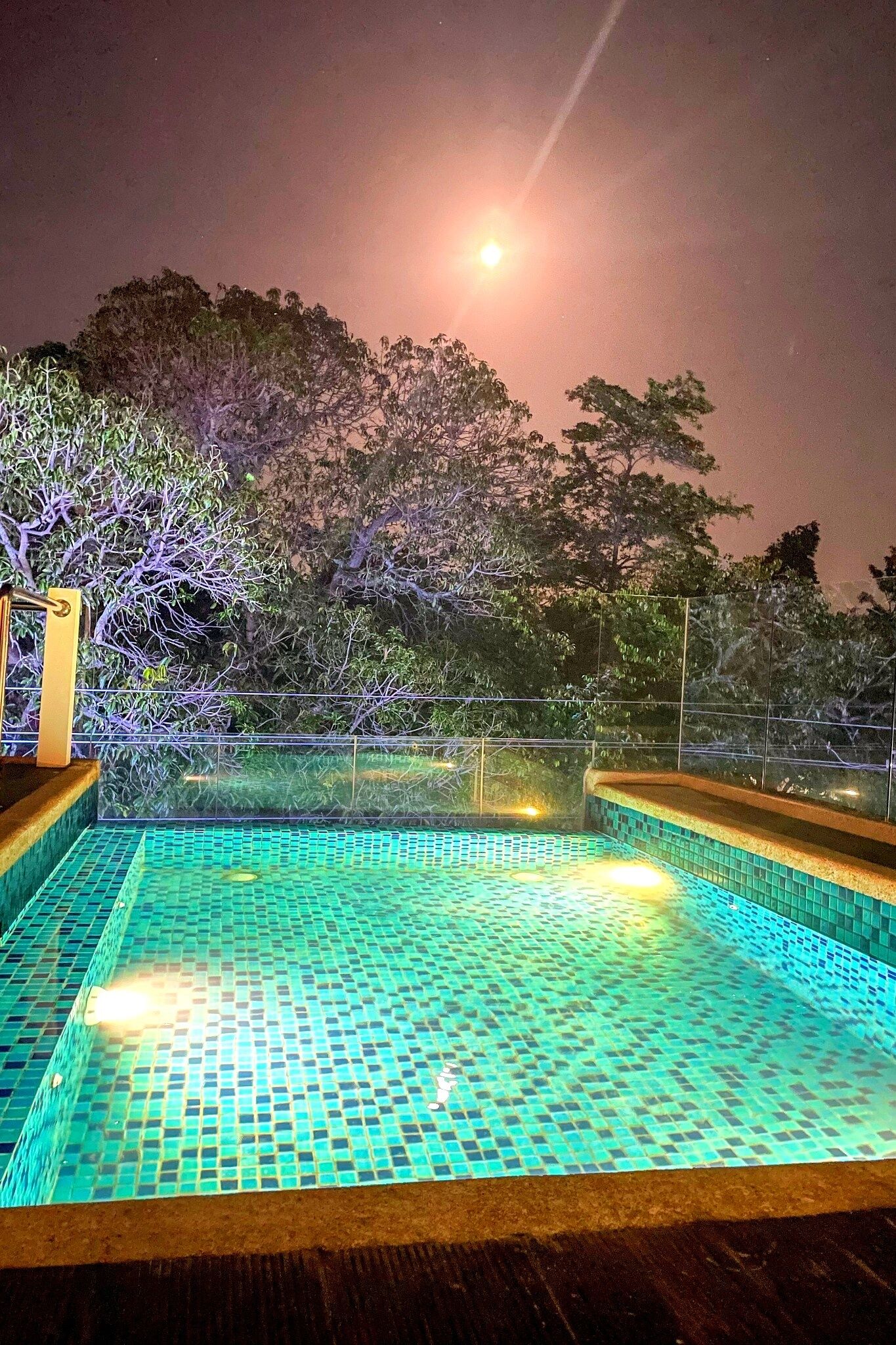 Uma área de piscina à noite com piso cerâmico, cercada por árvores e um muro.