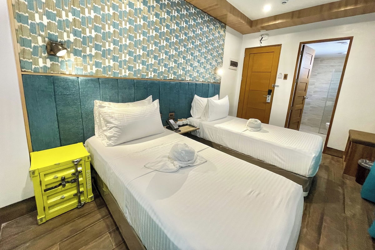 A hotel room with two beds, a bedside table, a wall clock, and a patterned wall.