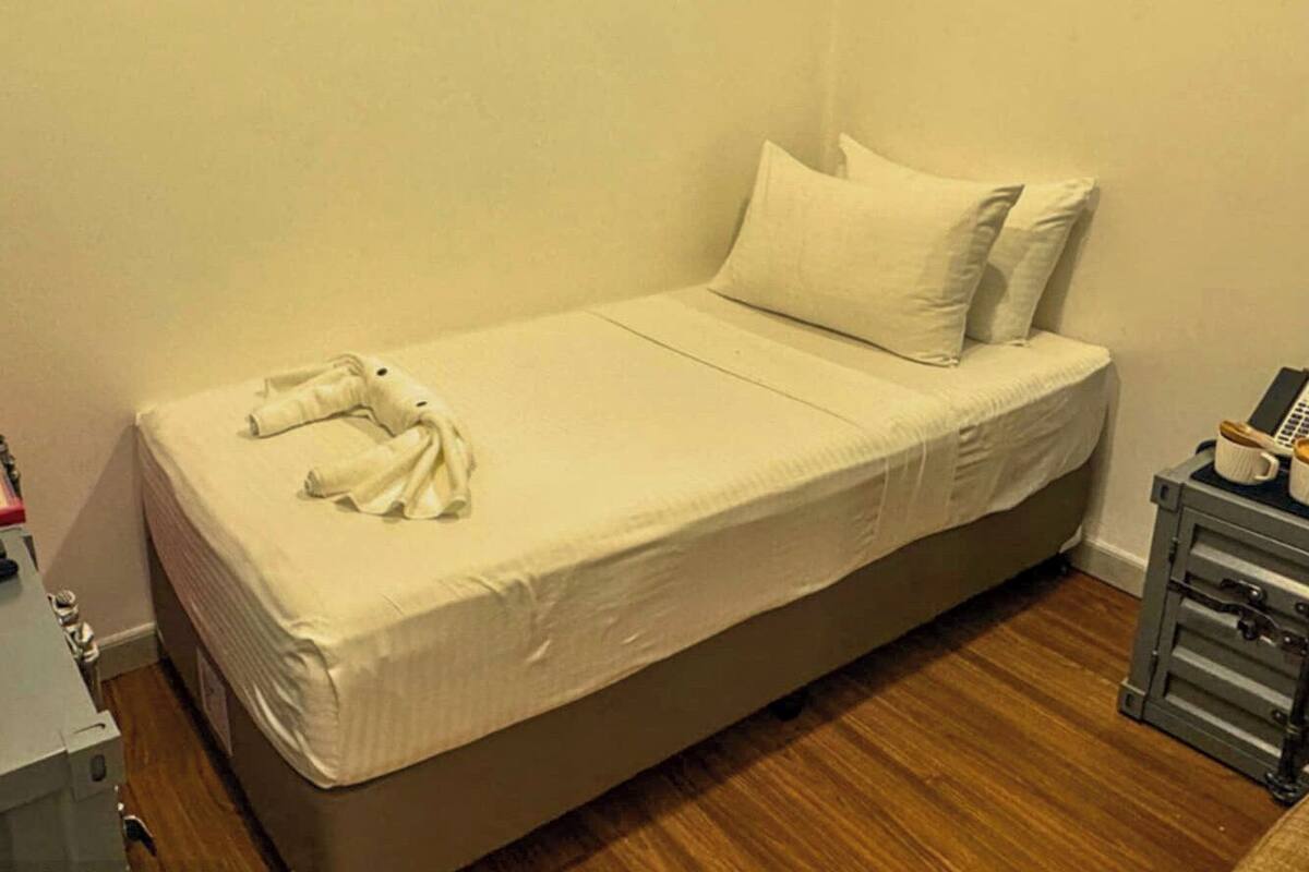 A single bed with a white mattress and pillows, a wooden floor, and a small bedside table with a phone and a cup.