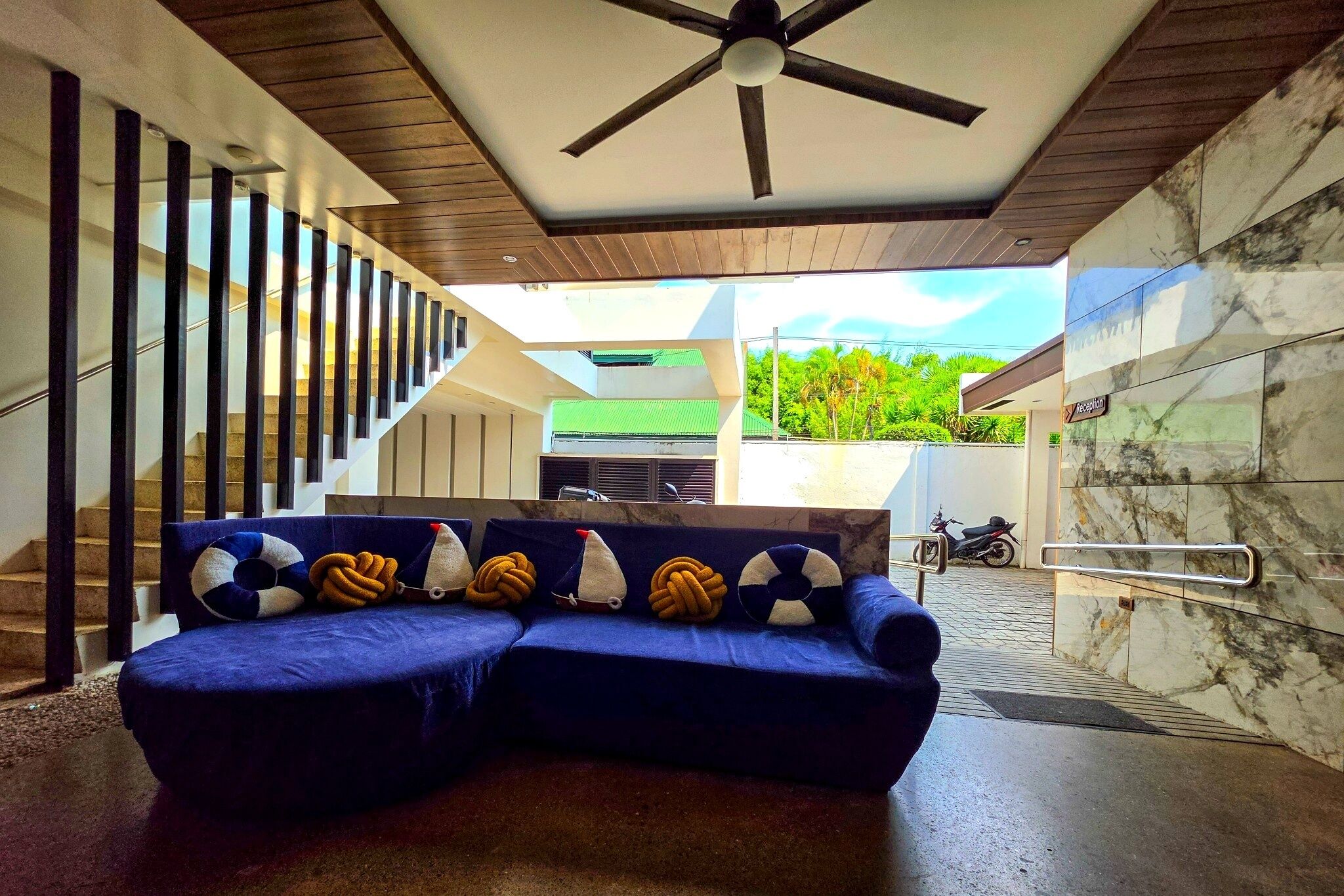 A modern interior with a blue sofa, a ceiling fan, a staircase, and a view of greenery outside.