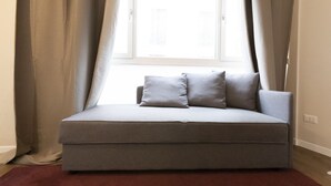 Een grijze bank met kussens in een kamer met een raam, gordijnen en een rood vloerkleed.