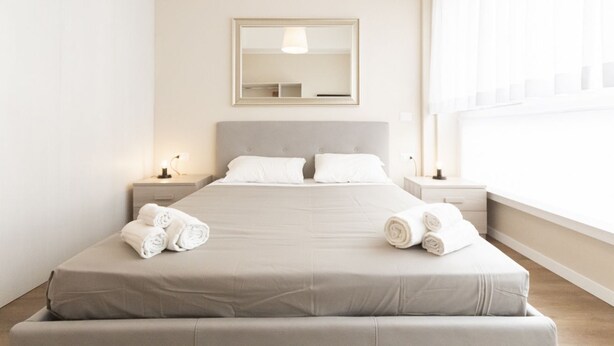 A neatly made bed with rolled towels, a bedside table with a lamp, and a large window with sheer curtains. - S.Maria Valle Apartment 3 (Milano)