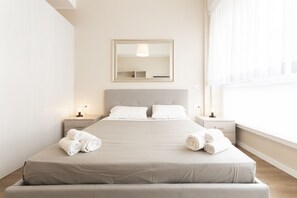 Ein ordentlich bezogenes Bett mit aufgerollten Handtüchern, ein Nachttisch mit Lampe und ein großes Fenster mit durchsichtigen Vorhängen.
