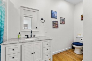 Ein Badezimmer mit weißem Waschtisch, einem Waschbecken, einem Spiegel und einer Toilette. An der Wand befinden sich Bilder in Rahmen.