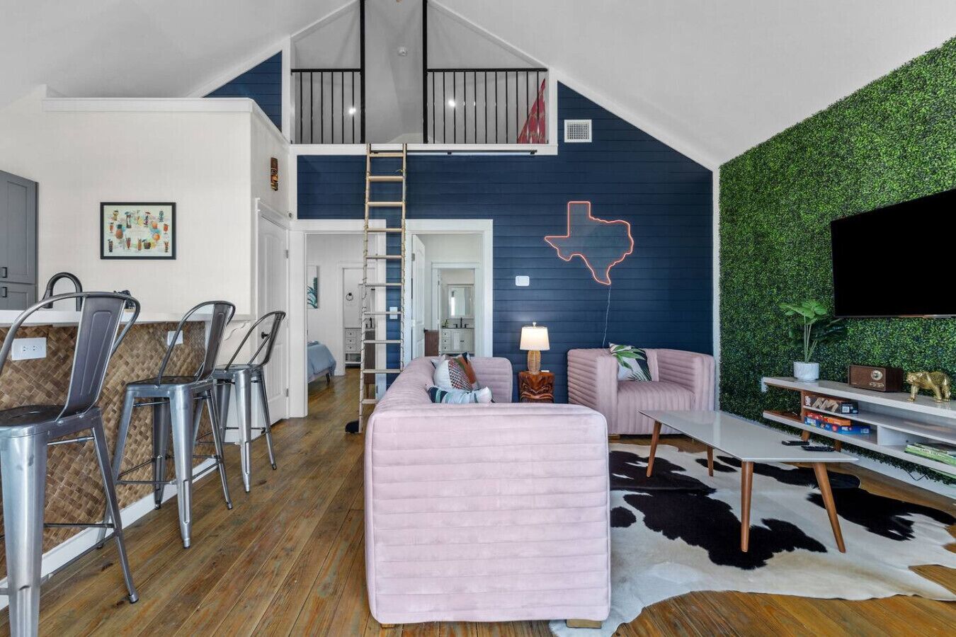 Un appartement moderne de style « loft » avec un lit en mezzanine, un coin repas avec des chaises en métal, un salon avec un canapé rose, un téléviseur fixé à un mur vert et un tapis en peau de vache.