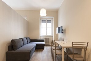 Un salon moderne comprenant un canapé d’angle, une table à manger avec des chaises, un téléviseur fixé au mur et une fenêtre avec des stores.