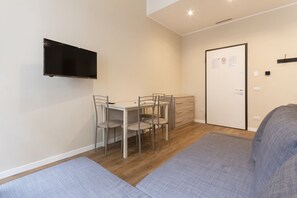 Une petite pièce avec un téléviseur fixé au mur, un bureau avec des chaises, une porte sur laquelle est affichée une note, et un canapé gris.