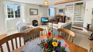 A cozy living room with a fireplace, a dining table with a floral centerpiece, and a staircase leading to an upper floor. - Orchard Cottage, Brayford (Brayford)