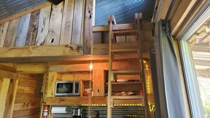 Een rustiek houten interieur met houten plafond, raam met metalen frame, een magnetron, een ladder en planken met servies.