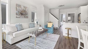A modern living room with a white sofa, a tufted ottoman, a blue armchair, a bar area with stools, and a staircase leading to another level.