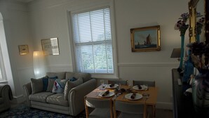 A living room with a sofa, a dining table set for two, and a window with blinds. - Lovely, spacious apartment - sleeps 5 (Ryde)