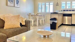 A modern living room with a glass coffee table, a sofa with a throw blanket, a dining area with white chairs, and a kitchen with stainless steel appliances. - 5 mins walk to town and beach. Family sized top floor condo! (Port Douglas)