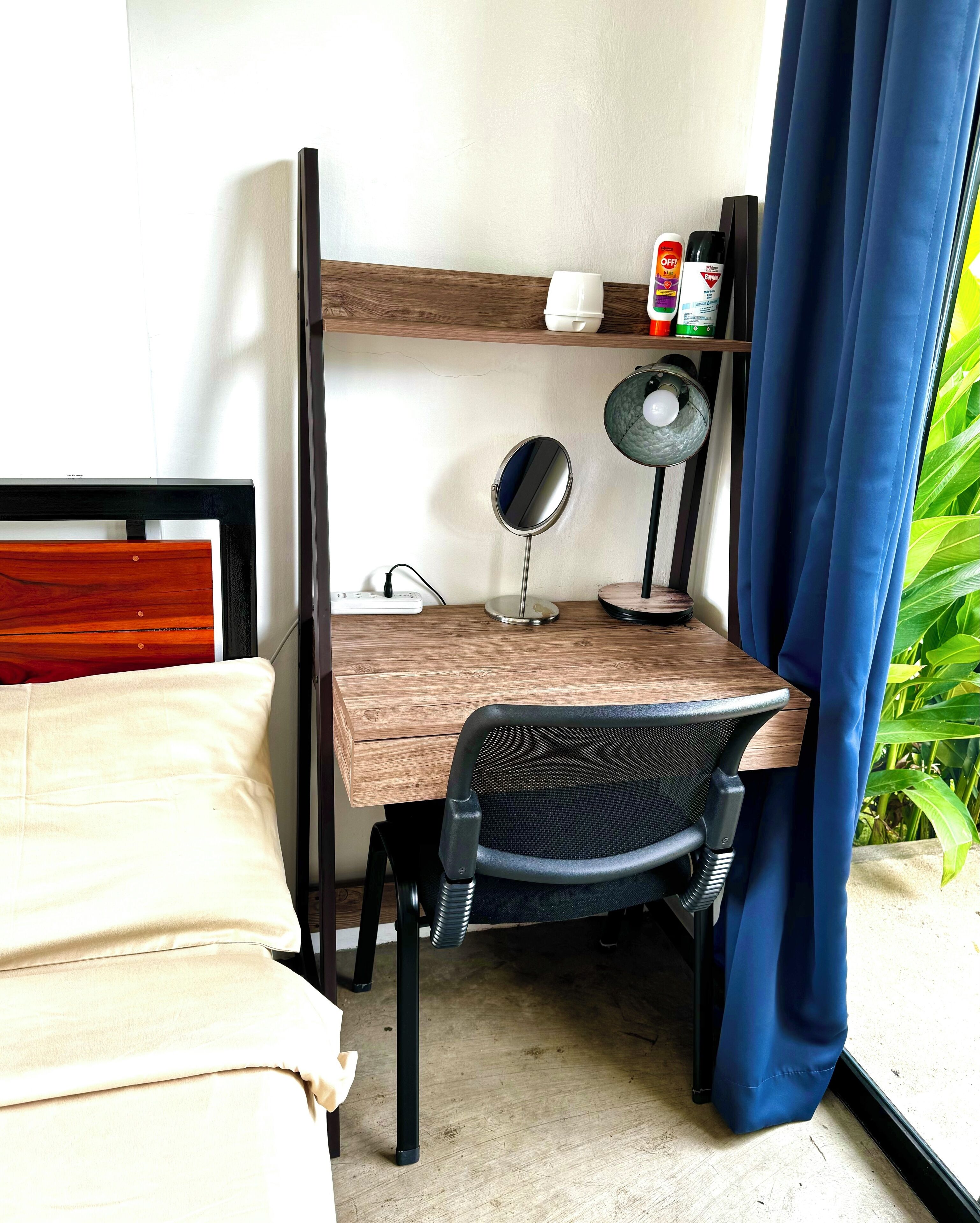 A compact room with a wooden desk, a black chair, a small mirror, and a shelf with a few items.