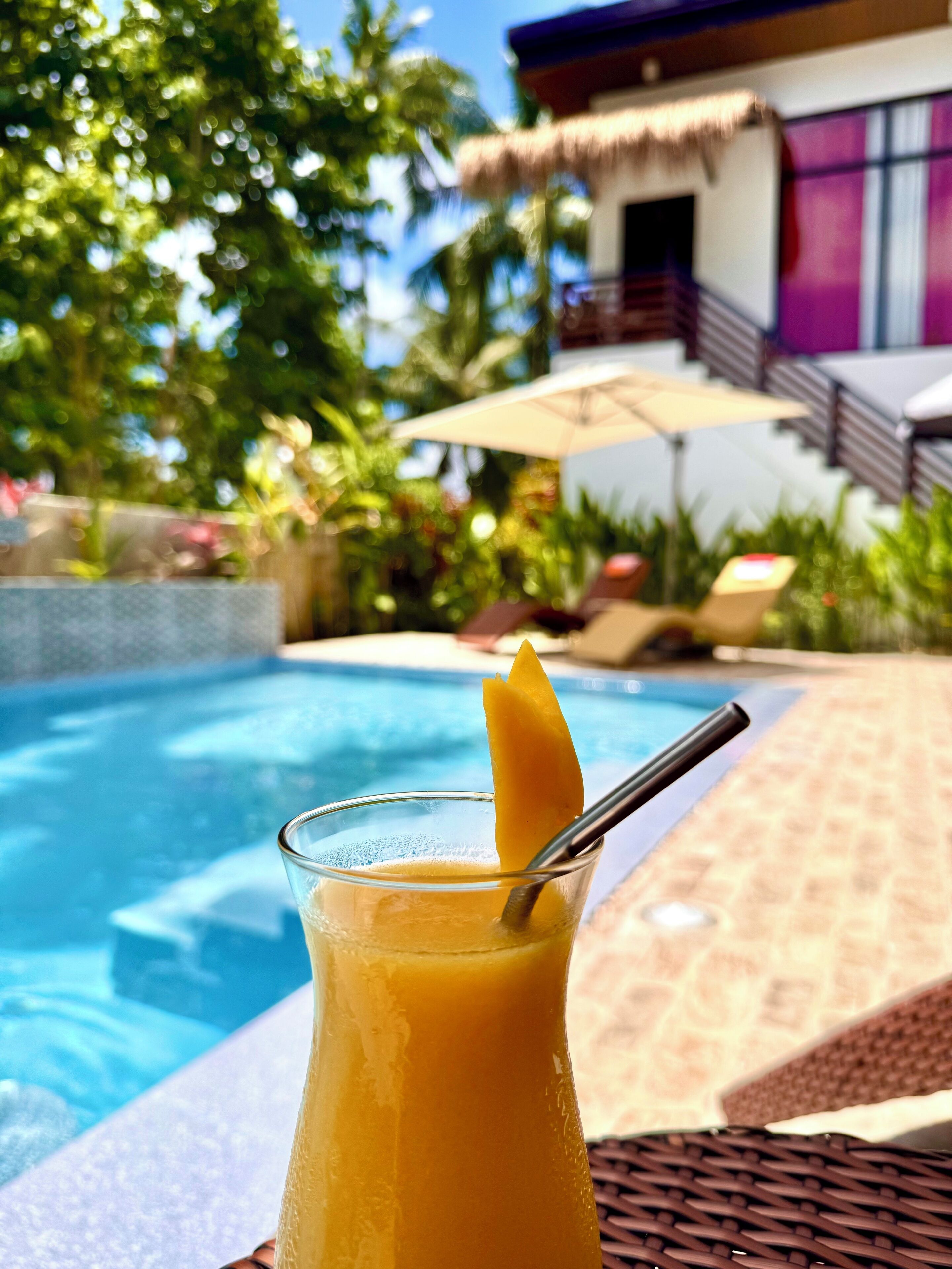 A glass of orange juice with a slice of orange and a straw, placed on a wicker chair by a poolside.