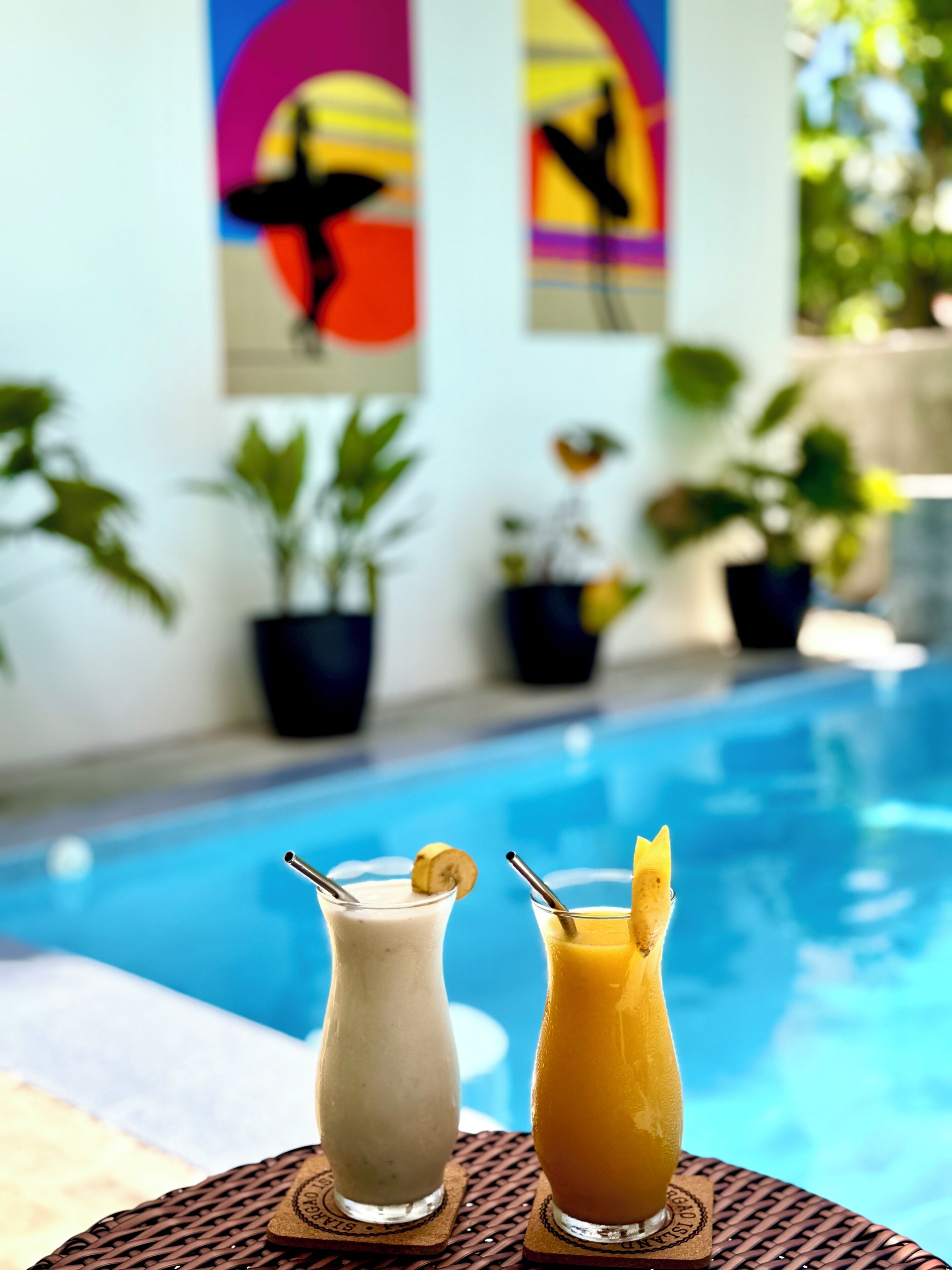 A poolside setting with two drinks on coasters, a pool, and potted plants.