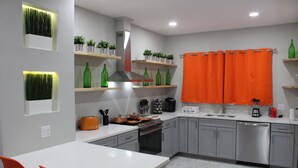 Une cuisine moderne avec des meubles gris, des plans de travail blancs et des touches de couleur orange. On y trouve des étagères avec des bouteilles vertes et des plantes, une cuisinière et un lave-vaisselle.