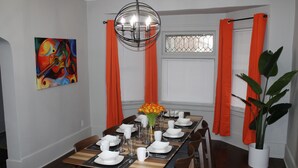Une salle à manger avec une table en bois dressée pour six personnes, entourée de chaises, d’un lustre et d’un tableau accroché au mur.
