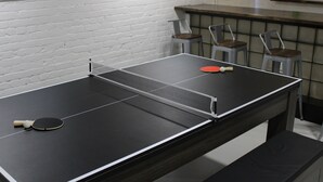 Une table de ping-pong avec deux raquettes et une balle, entourée de chaises en métal et d’un mur de briques.