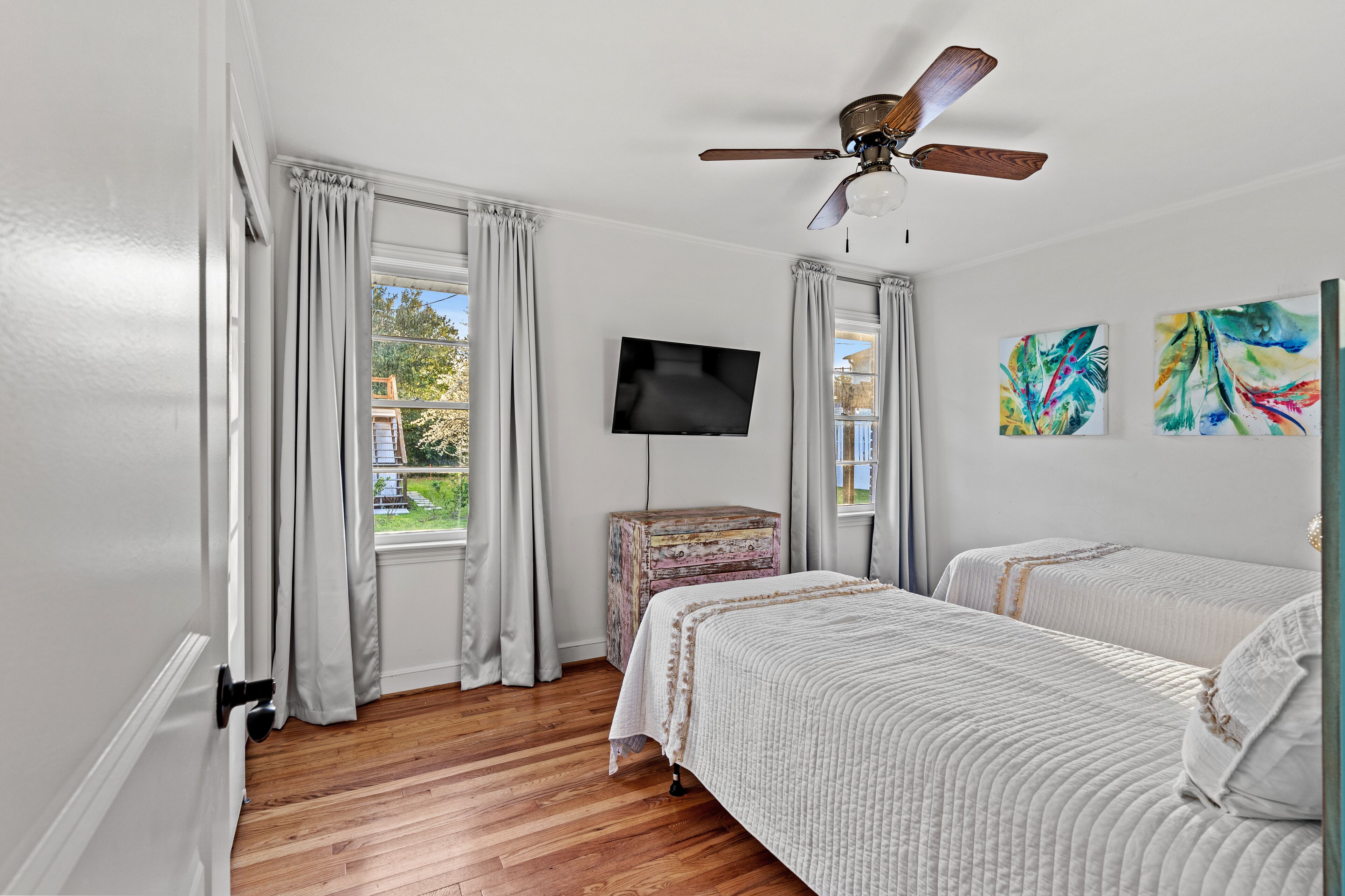 A bedroom with two beds, a ceiling fan, a TV, and artwork on the wall.