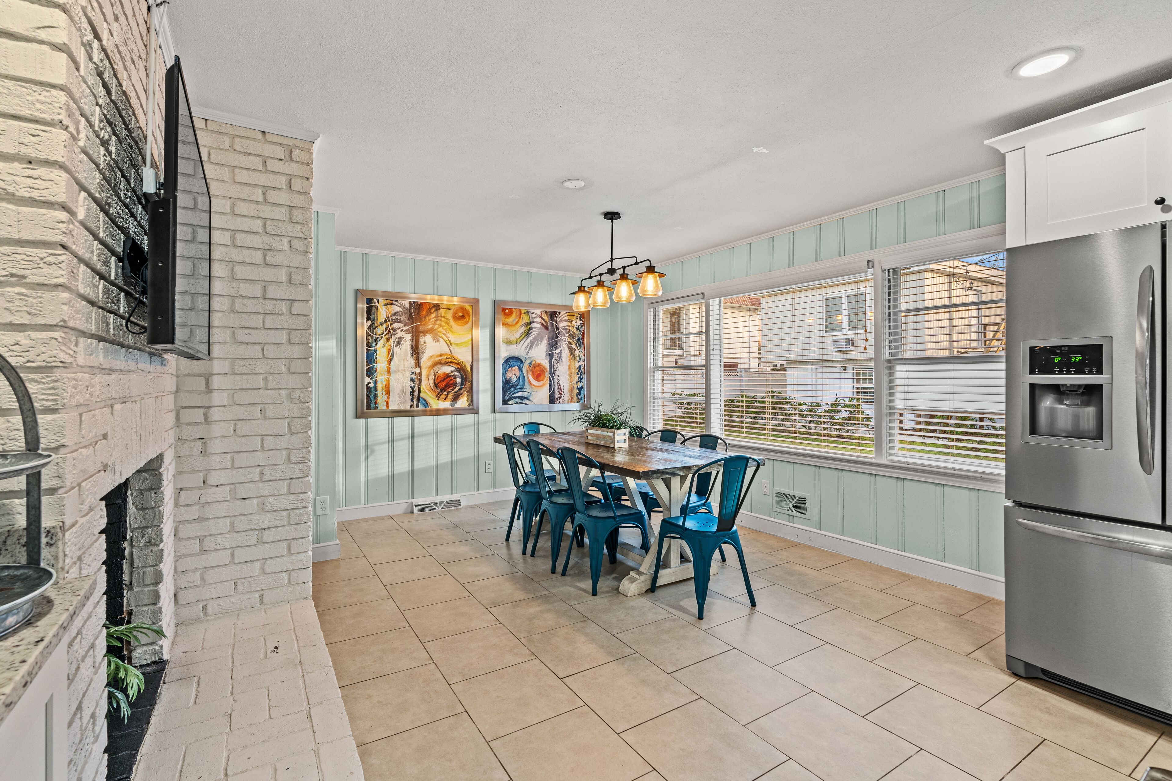 A kitchen with a brick wall, a dining table with chairs, a refrigerator, and a window with blinds.