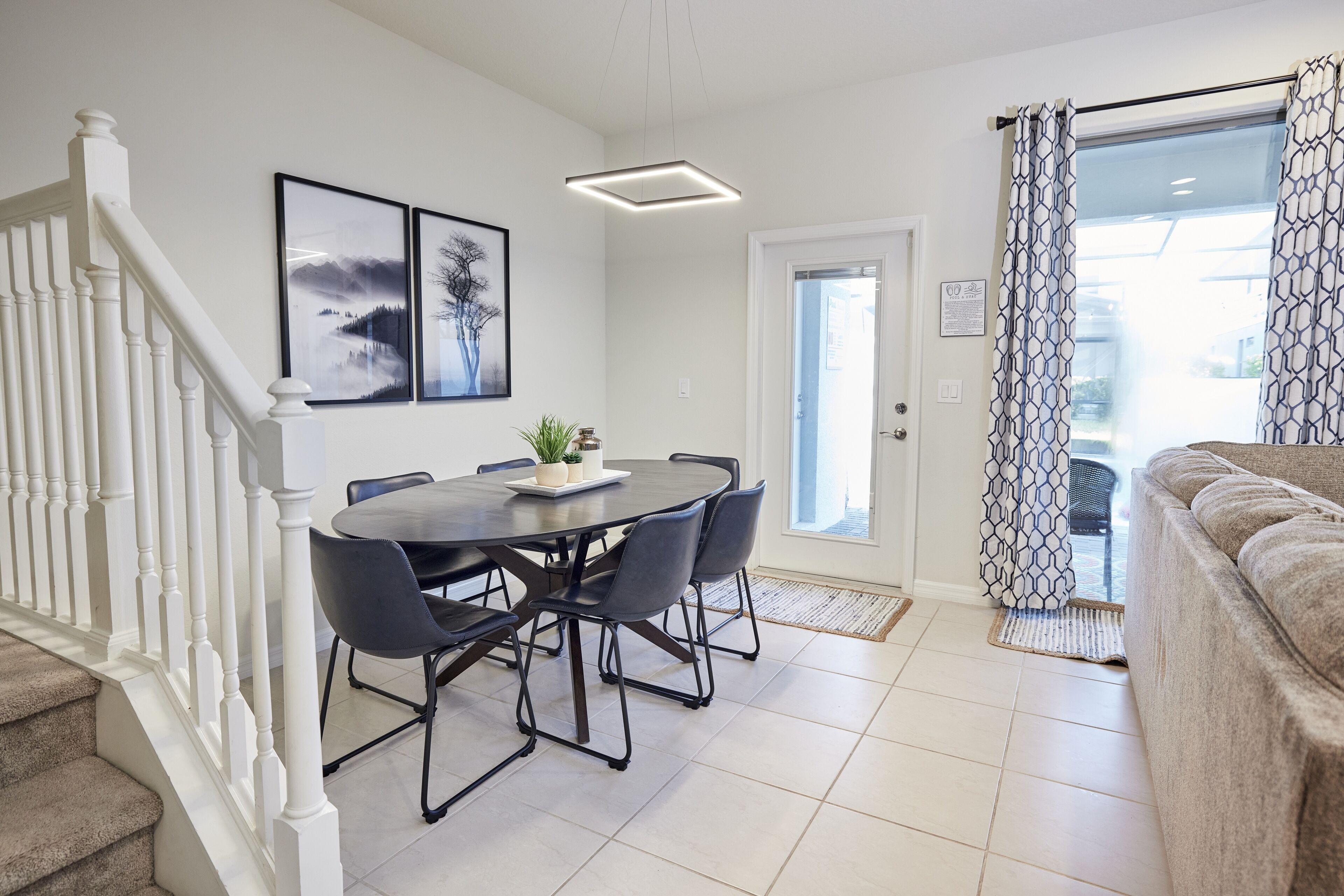 Una sala moderna con un sofá color beige, un área de comedor con una mesa redonda y sillas, una escalera con barandillas blancas y un gran ventanal con cortinas estampadas.