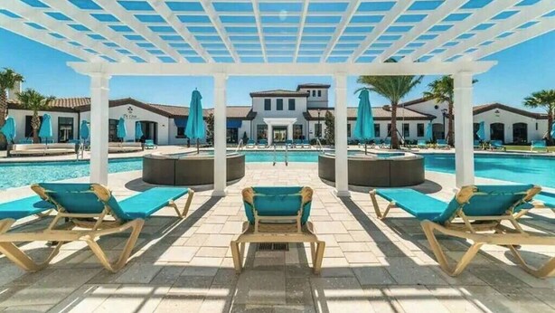 Un espace aménagé au bord de la piscine, avec des chaises longues et des parasols, un coin salon couvert et un bâtiment en arrière-plan.