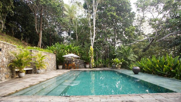 A rectangular swimming pool with a stone deck surrounding it, set in a lush garden with various plants and trees. - Quintessential colonial villa with pool and full staff (Habaraduwa)