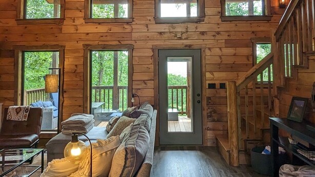 A cozy cabin interior with a wooden floor, a sofa, a table, and a staircase. - 3 BR Luxury Cabin & Hot Tub near Buffalo River! (Jasper)